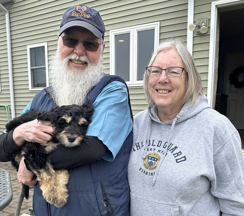 A couple smile at the camera. The man is wearing sunglasses, a baseball cap and has a long, full white beard and is holding a black and tan dog. The woman is wearing glasses and a gray hooded sweatshirt.